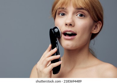 A Happy, Radiant Woman With Perfect Skin Stands On A Gray Background Wrapped In A Towel And Does A Facial Massage With A Black Electric Anti-aging Massager, Smiling Pleasantly At The Camera.