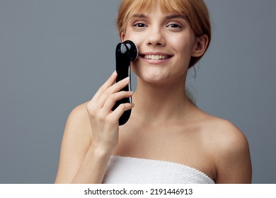 A Happy, Radiant Woman With Perfect Skin Stands On A Gray Background Wrapped In A Towel And Does A Facial Massage With A Black Electric Anti-aging Massager, Smiling Pleasantly At The Camera.