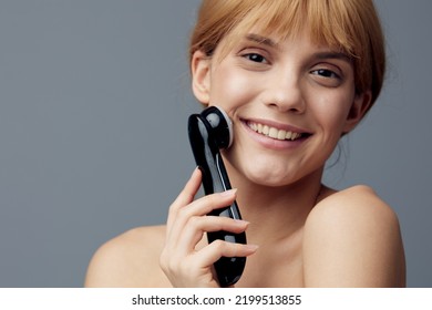 A Happy, Radiant Woman With Perfect Skin Stands On A Gray Background Wrapped In A Towel And Does A Facial Massage With A Black Electric Anti-aging Massager, Smiling Pleasantly At The Camera.