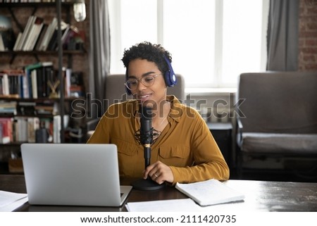 Happy radio channel newscaster, African journalist in headphones speaking at professional microphone, smiling at laptop display, presenting news, podcast, audio post on air, using sound equipment