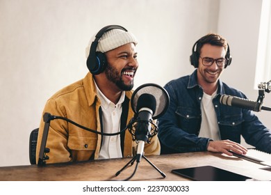 Happy radio presenter smiling while speaking into a microphone in a studio. Cheerful young man co-hosting an audio broadcast with a guest. Two young content creators recording an internet podcast.