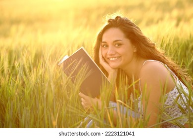 Happy Reader Holding Book Looks At Camera In A Wheat Field At Sunset