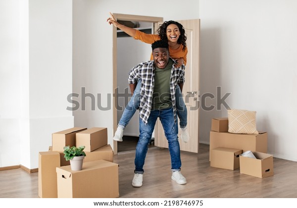 Happy Real Estate\
Buyers Couple Celebrating Moving New House. African American\
Husband Carrying Wife Piggyback Having Fun Posing In Rented\
Apartment, Smiling To\
Camera