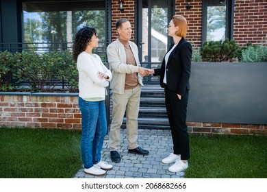 happy realtor in glasses shaking hands with smiling man standing near african american wife and modern house