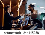 Happy receptionist giving room cardkey to African American hotel guest at reception desk.