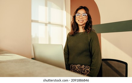 Happy Receptionist Standing At The Front Desk Of A Co-working Space. Friendly Female Assistant Smiling At The Camera Cheerfully While Working At The Reception In A Modern Office.