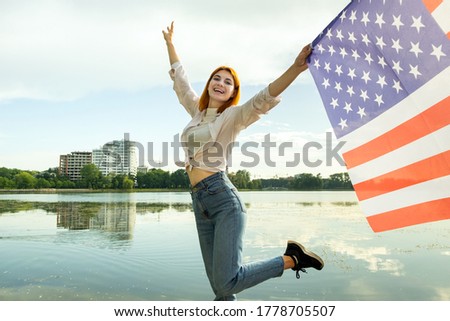 Happy red haired young woman with United States national flag in her hand. Positive girl celebrating US independence day.