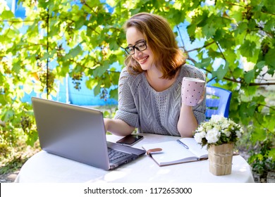 A happy red-haired woman blogger in black glasses is sitting outside in the garden and working online. Modern concept of freelancing, digital nomad, remote work. - Shutterstock ID 1172653000