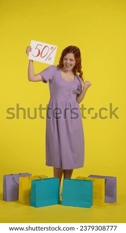 Happy redhaired woman surrounded by shopping bags holding a poster with the inscription fifty percent. A woman makes a victory gesture with her hand in the studio on a yellow background. Vertical shot