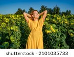 A happy red-haired woman in a yellow dress smiles with her eyes closed, exposing her face to the sun in a sunflower field