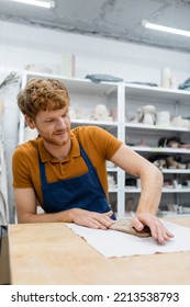 Happy Redhead Man In Apron Pressing Clay Piece With Hand During Pottery Class