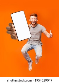Happy redhead man jumping up in air, showing thumbs up gesture, demonstrating smartphone with empty screen, recommending new mobile application on orange studio background - Shutterstock ID 1954329049