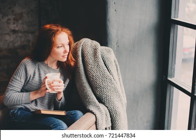 Happy Redhead Woman Relaxing At Home In Cozy Winter Or Autumn Weekend With Book And Cup Of Hot Tea, Sitting In Soft Chair With Blanket