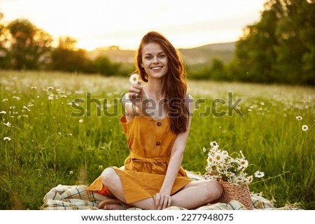 happy redhead woman sitting in a chamomile field on a plaid in a lotus position and holding a flower in her hands smiling at the camera