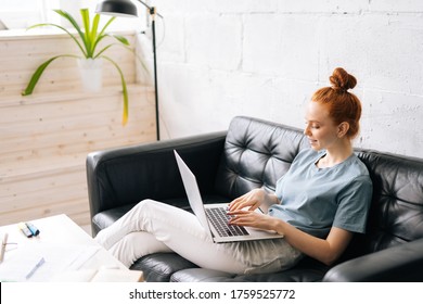 Happy redhead young woman using laptop at home sitting on soft couch. Lady is working on laptop computer. Girl student is typing message using laptop keyboard