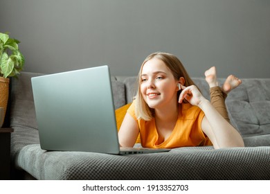 Happy relax young woman lies on sofa with laptop in headphones. Remote work, online study, online shopping watching films or video call on laptop. Teen girl rest at home interior of living room