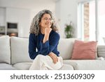 Happy relaxed 50 years old woman sitting on sofa at home looking away. Smiling middle aged lady posing in living room at home. Mature older female with curly gray hair relaxing on couch enjoying chill