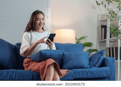 Happy and relaxed Asian woman sitting on sofa on weekend using smartphone to communicate, talk, text, chat online, watch wireless internet on device. Shopping online from your home office. - Powered by Shutterstock