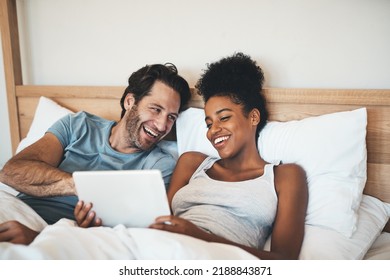 Happy, Relaxed And Carefree Couple Reading Social Media News On Digital Tablet And Laughing In Bed. Interracial Husband And Wife Waking Up Together And Browsing Internet, Sharing A Funny Online Joke