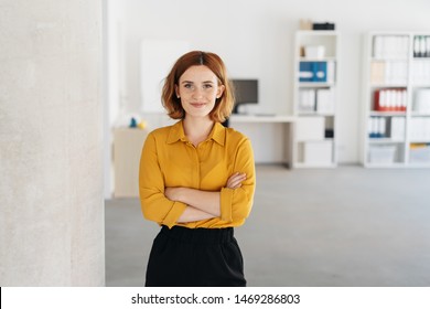 Feliz, relaxado, confiante jovem empresária de pé com os braços cruzados em um escritório espaçoso, olhando para a câmera com um sorriso caloroso e amigável