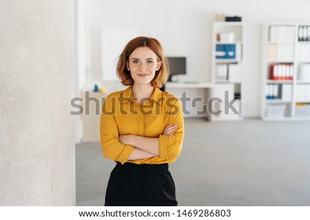 Happy relaxed confident young businesswoman standing with folded arms in a spacious office looking at the camera with a warm friendly smile
