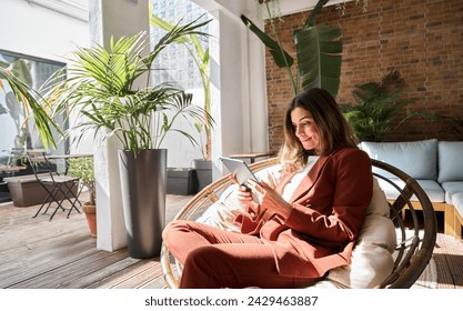 Happy relaxed elegant mature middle aged professional business woman executive wearing suit holding digital tablet using tab working sitting in comfortable chair in sunny office with green plants. - Powered by Shutterstock