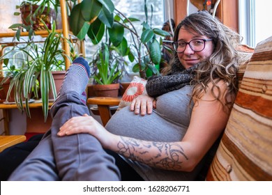 Happy And Relaxed Expectant Parents Sit Together In Living Room, First Person Perspective As Man Rests Legs In Lap Of Heavily Pregnant Woman