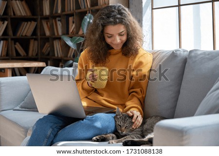 Happy relaxed hispanic teen girl student or freelancer relaxing sitting on couch holding laptop on lap working studying from home drinking tea, playing with cute pet cat in cozy sunny room on sofa.