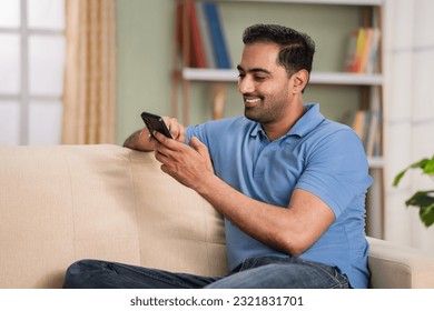 Happy relaxed indian man using mobile phone while sitting on sofa at home during sunday - concept of online dating, relaxation and social media.
