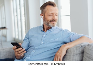 Happy relaxed man with a blissful smile sitting listening to music on his mobile phone using an earbud as he sits on a sofa at home