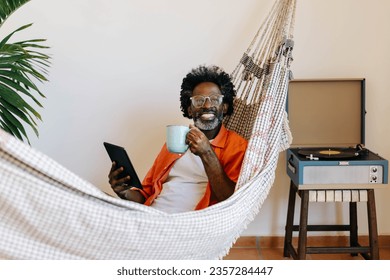 Happy, relaxed man holds a cup of Brazilian coffee while sitting in a hammock. Mature black man browsing the internet on a digital tablet, enjoying a leisurely moment at home. - Powered by Shutterstock