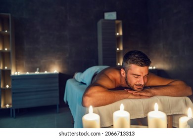 A happy relaxed man lying down on a massage table at the spa center and enjoying peaceful moments surrounded by candles. Massage and aromatherapy. The man at massage in spa center.