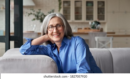 Happy relaxed mature old adult woman wearing glasses resting sitting on couch at home. Smiling middle aged grey-haired elegant senior lady relaxing on comfortable sofa looking at camera. Portrait.