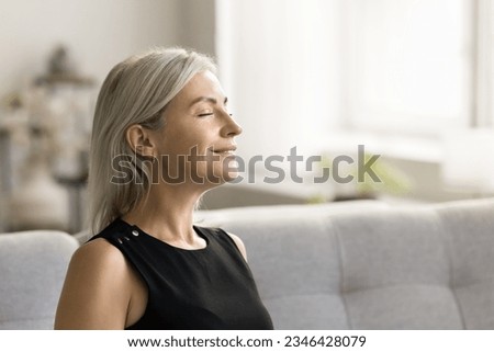 Happy relaxed mature woman enjoying fresh air, smells, relaxation, leisure at home, taking deep breath with closed eyes, smiling, meditating, reloading mind, enjoying stress relief technique