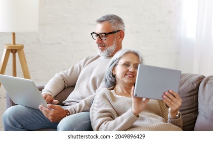 Happy relaxed old retired family couple using modern technologies laptop and digital tablet while relaxing resting on sofa in living room, senior man and woman surfing internet in free time