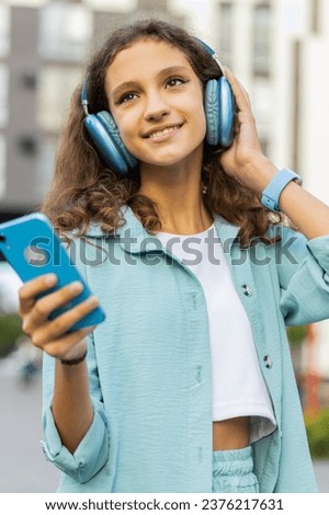 Happy relaxed overjoyed preteen child kid in wireless headphones choosing, listening favorite energetic disco rock n roll music dancing outdoors. Girl walking in urban european city street background