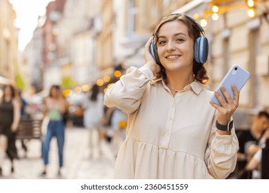 Happy relaxed overjoyed pretty young woman in wireless headphones choosing, listening favorite energetic disco rock n roll music dancing outdoors. Girl walking in urban city sunshine street background - Powered by Shutterstock