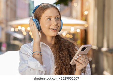 Happy relaxed overjoyed teenager girl in wireless headphones choosing, listening favorite energetic disco music in smartphone dancing outdoors. Child tourist walking passes by urban sunny city street - Powered by Shutterstock