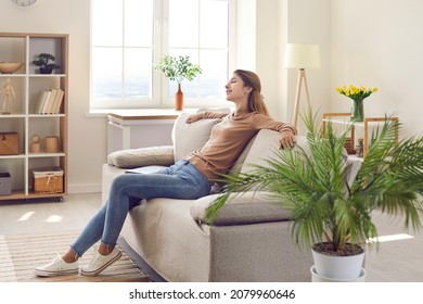 Happy relaxed woman resting on comfy couch in modern interior of her own apartment. Young girl spending free time at home, enjoying quiet leisure, sitting on cosy sofa in living room. Comfort concept