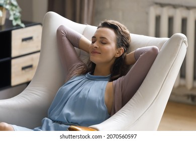 Happy relaxed young 30s Caucasian woman daydreaming in cozy armchair, sleeping napping seeing sweet dreams, enjoying carefree weekend leisure time in modern living room, meditating alone at home.