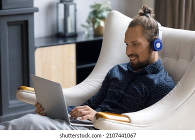 Happy Relaxed Young Handsome Man In Wireless Headphones Looking At Laptop Screen, Enjoying Distant Video Call Conversation Or Watching Funny Comedian Movie Online, Sitting On Cozy Armchair At Home.