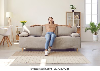 Happy relaxed young woman resting sitting on comfortable sofa in living room at home. Portrait of girl in casual clothes enjoying lazy weekend or vacation relaxing in cozy room of her home.