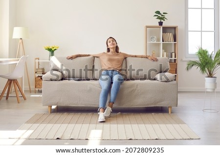 Happy relaxed young woman resting sitting on comfortable sofa in living room at home. Portrait of girl in casual clothes enjoying lazy weekend or vacation relaxing in cozy room of her home.