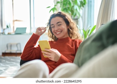 Feliz y relajada joven sentada en el sofá usando el teléfono celular, una mujer sonriente riendo sosteniendo el smartphone, mirando el teléfono celular disfrutando haciendo compras en línea por ecommerce en aplicaciones móviles o viendo videos.