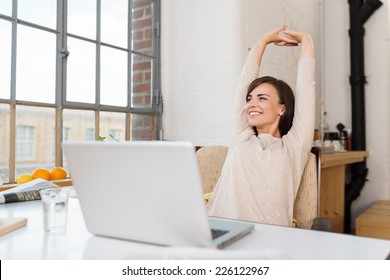 Happy relaxed young woman sitting in her kitchen with a laptop in front of her stretching her arms above her head and looking out of the window with a smile