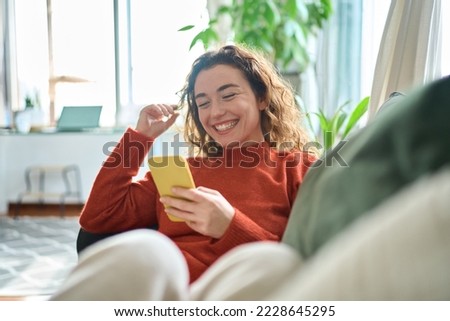 Photo of Happy relaxed young woman sitting on couch using cell phone, smiling lady laughing holding smartphone, looking at cellphone enjoying doing online ecommerce shopping in mobile apps or watching videos.