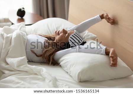 Happy relaxed young woman with sleeping mask awaken in cozy bedroom, stretching hands, feeling energetic. Smiling millennial mixed race girl enjoying lazy weekend morning in comfortable bed at home.