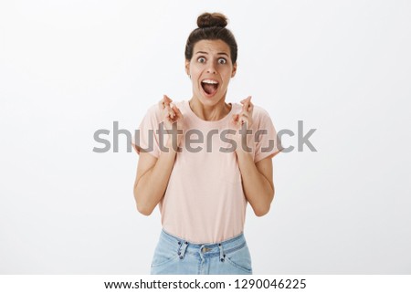 Happy relieved and joyful young woman smiling broadly feeling excited as pray worked standing with crossed fingers for good luck and seing she won award posing satisfied and joyful over grey wall