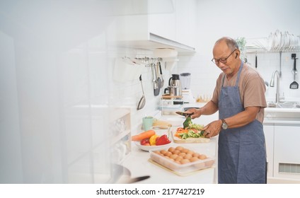 Happy Retired Asian Mature Adult Man Standing Chopping Fresh Broccoli On Wooden Board While Preparing Healthy Food, Senior Male Cooking In Modern Kitchen.