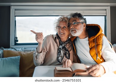 Happy retired couple enjoying their adventurous road trip, pointing at scenic view outside window of their cozy camper van while reading a guidebook - Powered by Shutterstock